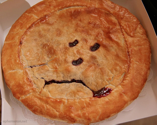 Photograph of a cherry pie in a bakery box, its crust cut with two eyes and a downturned mouth so that it looks like a sad face