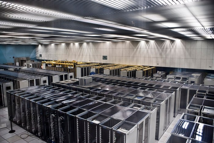 Photograph of a large data centre: rows of server racks receding under a ribbed metal ceiling