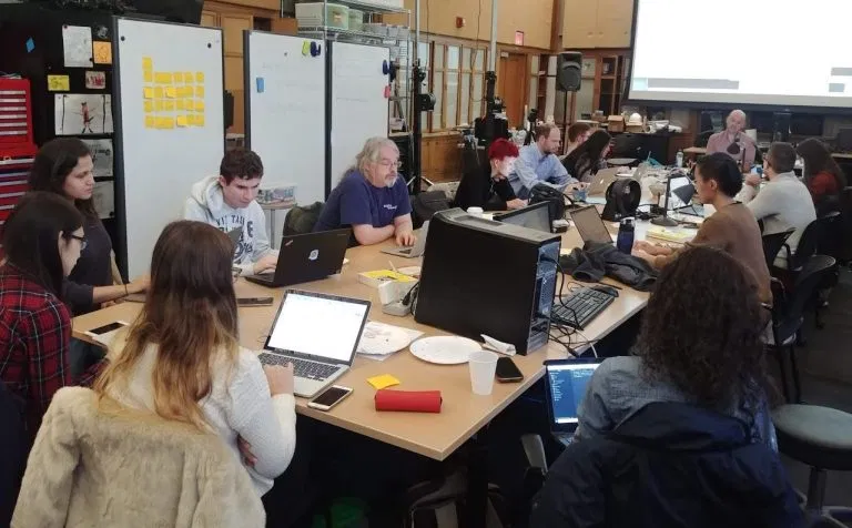 Participants around tables of laptops in the STUDIO for Creative Inquiry, with a wall of yellow post-it notes behind them.
