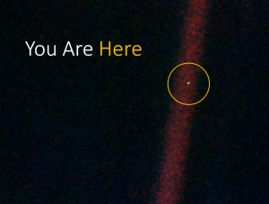 Photograph of the Voyager 1 “Pale Blue Dot”: Earth as a single bright speck in a band of scattered sunlight, captioned “You Are Here” with the speck circled in yellow