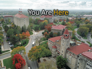 Aerial photograph of a university campus in autumn — brick and limestone buildings, a clock tower and tree-lined streets — captioned “You Are Here”