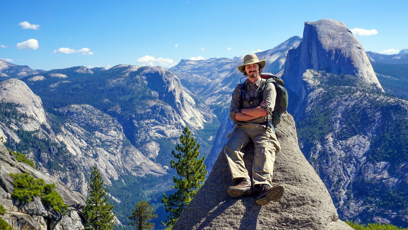 Scott Weingart in a sun hat and backpack, sitting on a granite outcrop at Glacier Point with Half Dome and Yosemite Valley behind him